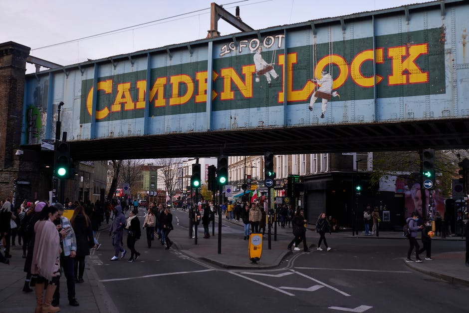 A busy street scene in Camden Town with a large, elevated metal railway bridge overhead, painted in weathered blue with bold yellow and red lettering spelling 'CAMDEN LOCK.' Under the bridge, numerous