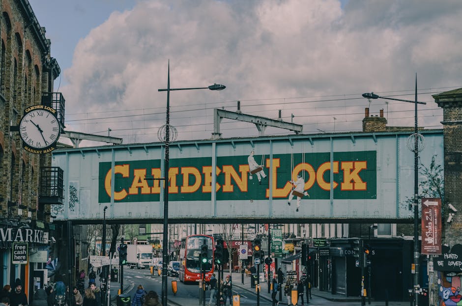 A busy street scene in Camden Town with a large, elevated metal railway bridge overhead, painted in weathered blue with bold yellow and red lettering spelling 'CAMDEN LOCK.' Under the bridge, numerous