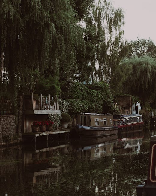 A tranquil scene along Regents Canal featuring two narrowboats moored close to a lush, green garden with overhanging trees, including weeping willows and dense foliage. The canal water reflects the bo
