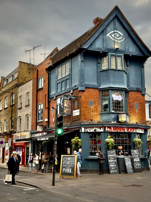 A multi-story building on a city street corner, featuring a blue-painted upper facade with decorative trim and small bay windows. The ground level houses a pub with large windows, flower boxes with gr